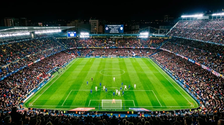 Estadio de fútbol inglés iluminado durante un partido nocturno con el césped intensamente verde